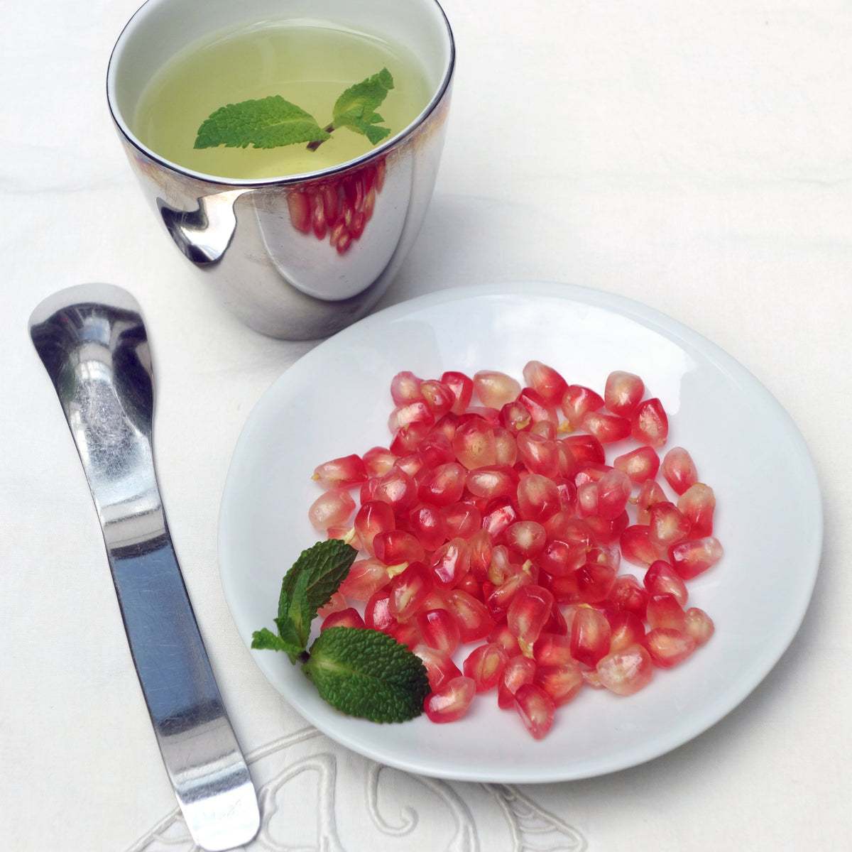 cuillère à dessert de forme irréulière, thé glacé dans une tasse argentée et grans de grenade et menthe dans une soucoupe blanche - tsé tsé