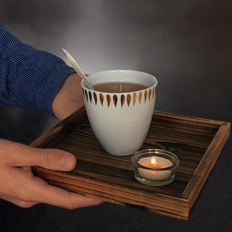 zoom sur mains tenant un plateau en bois avec tasse à thé en porcelaine blanche et or - tsé tsé