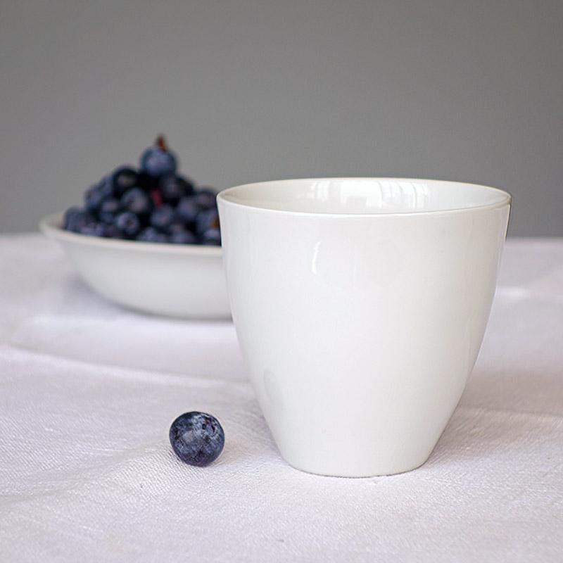 tasse blanche en porcelaine de forme irrégulière à côté d'une myrtille sur une nappe blanche - tsé tsé