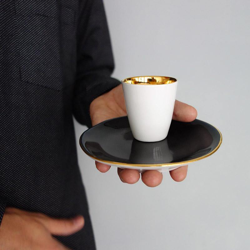 petite tasse à café en porelaine blanche intérieur or et soucoupe bleu nuit avec un liseré doré tenus en main - tsé tsé